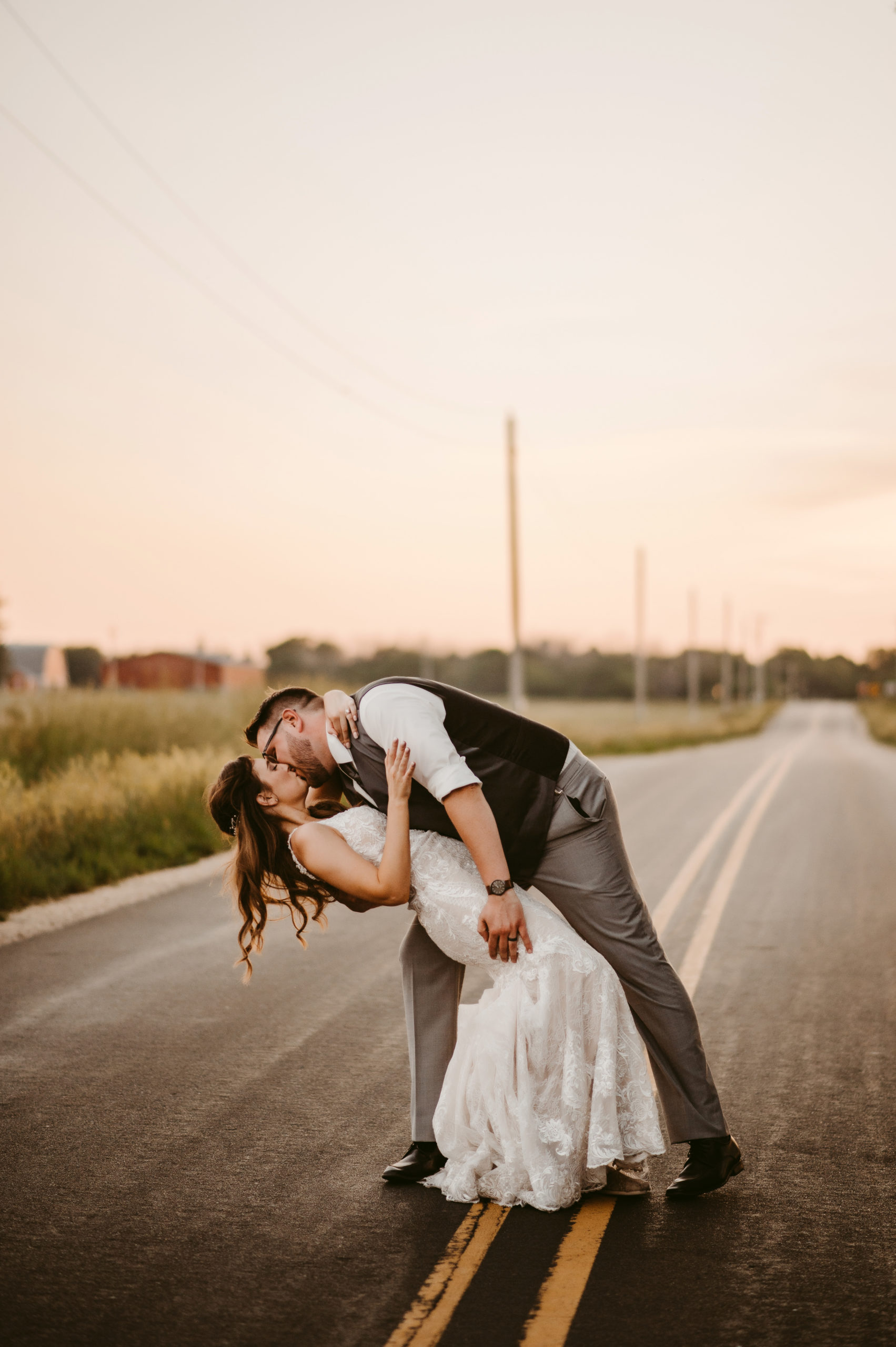 couple in wedding attire standing on a rural road at sunset, man dipping woman and kissing